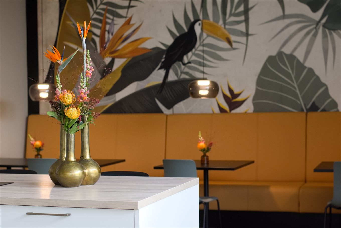 Goldfarbene Vasen mit Strelitzien vor einer senfgelben Sitzbank und botanischer Wandgestaltung.